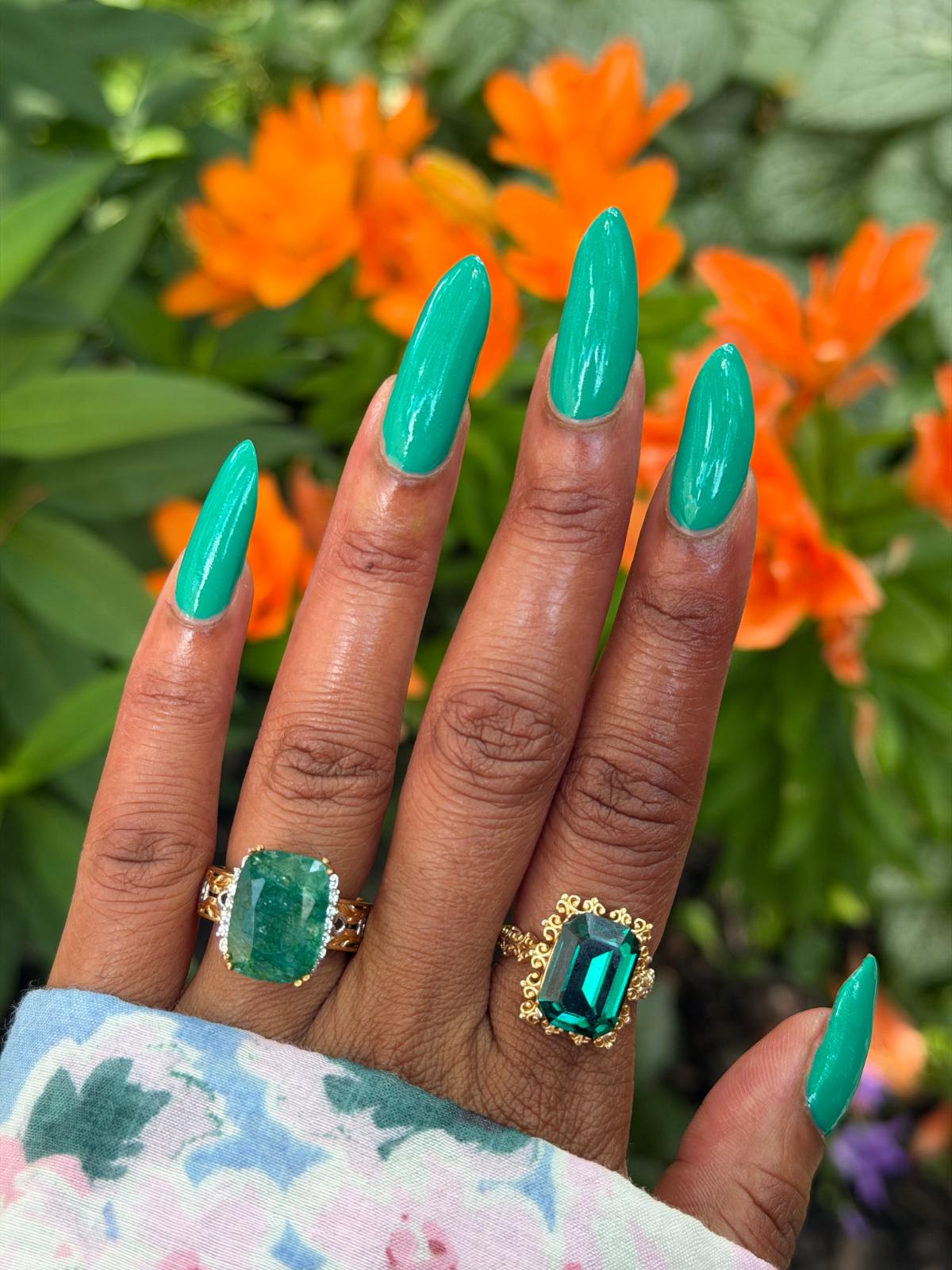 A close-up of a hand with long, glossy emerald green manicured nails, wearing two gold rings with large green gemstones, set against a blurred background of orange flowers and green leaves.