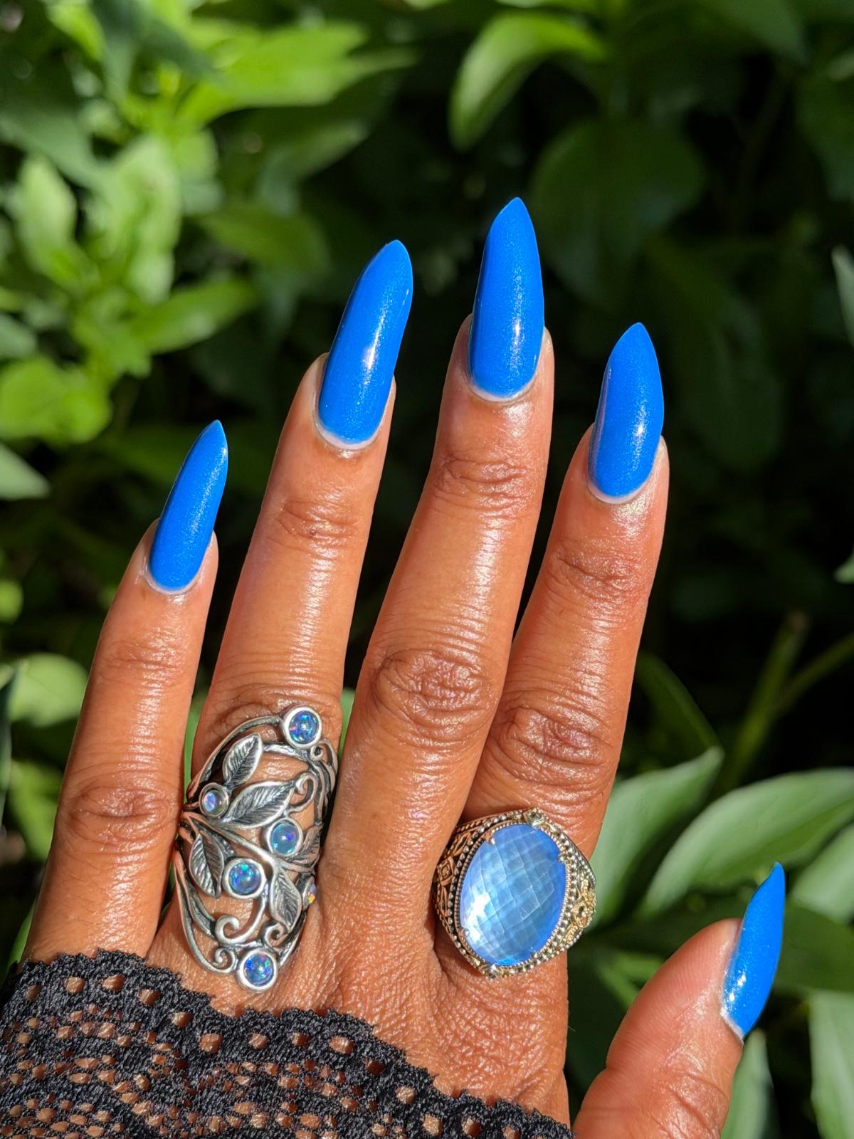 A close-up of a hand with bright blue manicured nails wearing two statement rings against a blurred green foliage background. One ring is a silver-toned leaf cluster design with blue stones, and the other is a large oval light blue stone ring.