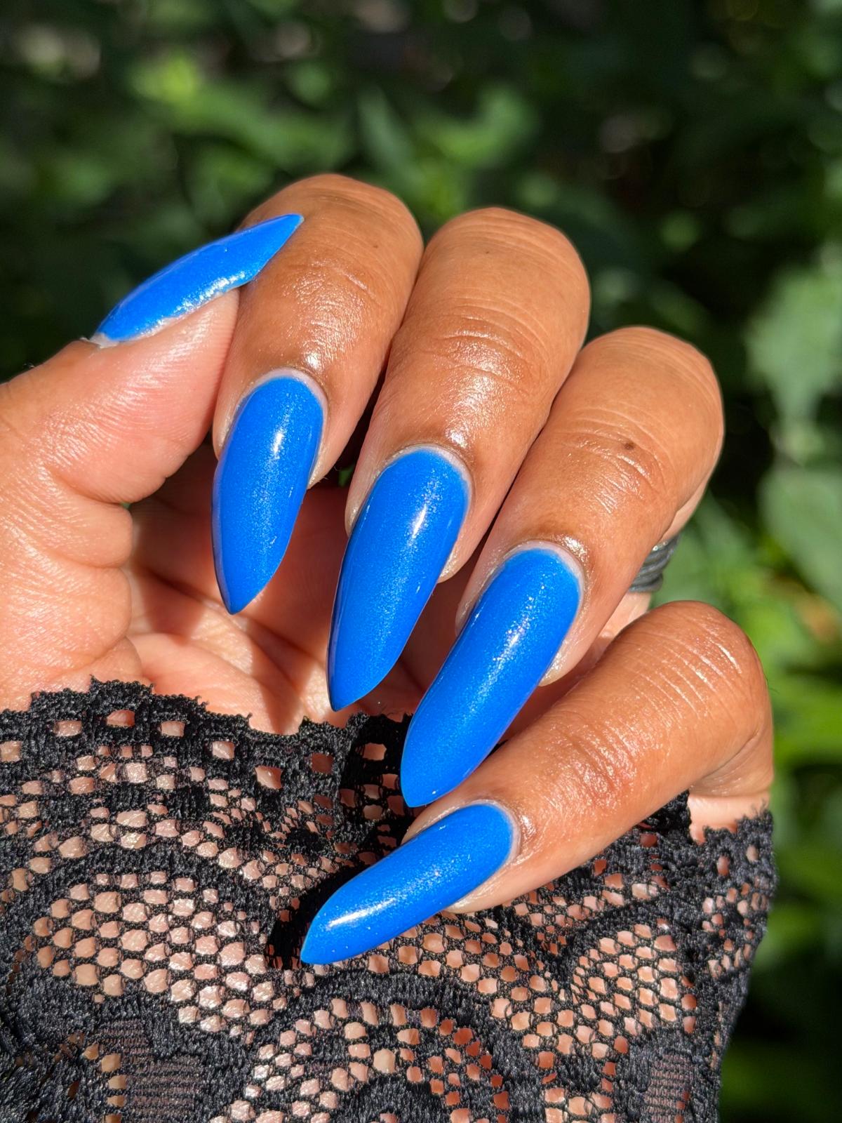 A close-up of a hand with long, glossy, bright blue stiletto-shaped nails, wearing a black lace sleeve, set against a blurred green outdoor background.