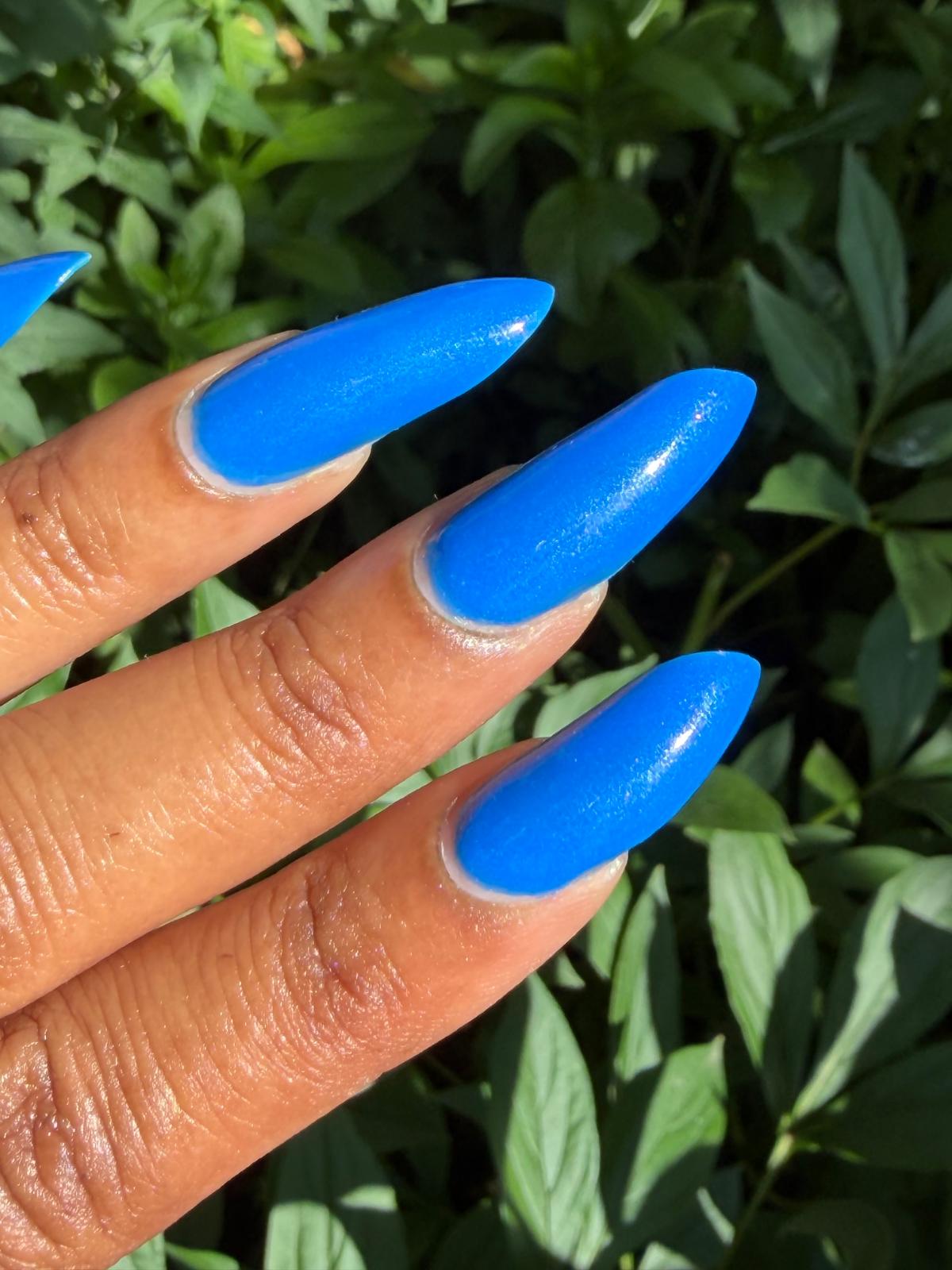 A close-up of a hand with long, glossy, bright blue stiletto-shaped nails, set against a background of green outdoor foliage.
