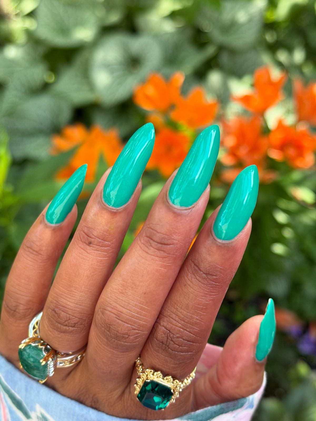 A close-up of a hand with long, glossy emerald green stiletto-shaped press-on nails, wearing gold rings with green gemstones, set against a blurred background of green foliage and orange flowers.
