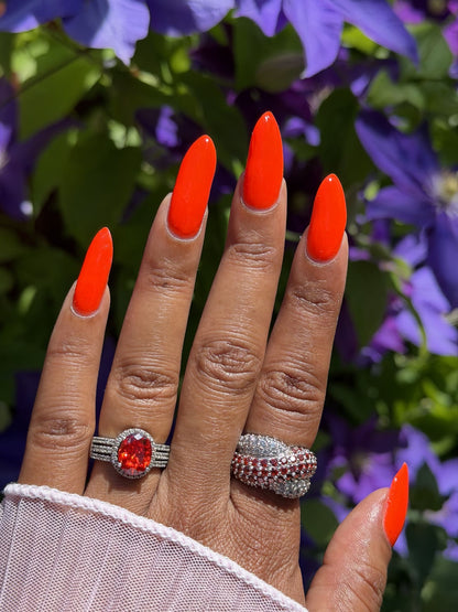 A close-up of a hand with bright orange manicured nails, wearing two silver rings: one with a large round red gemstone and another textured silver ring, set against a blurred background of purple flowers and green leaves.