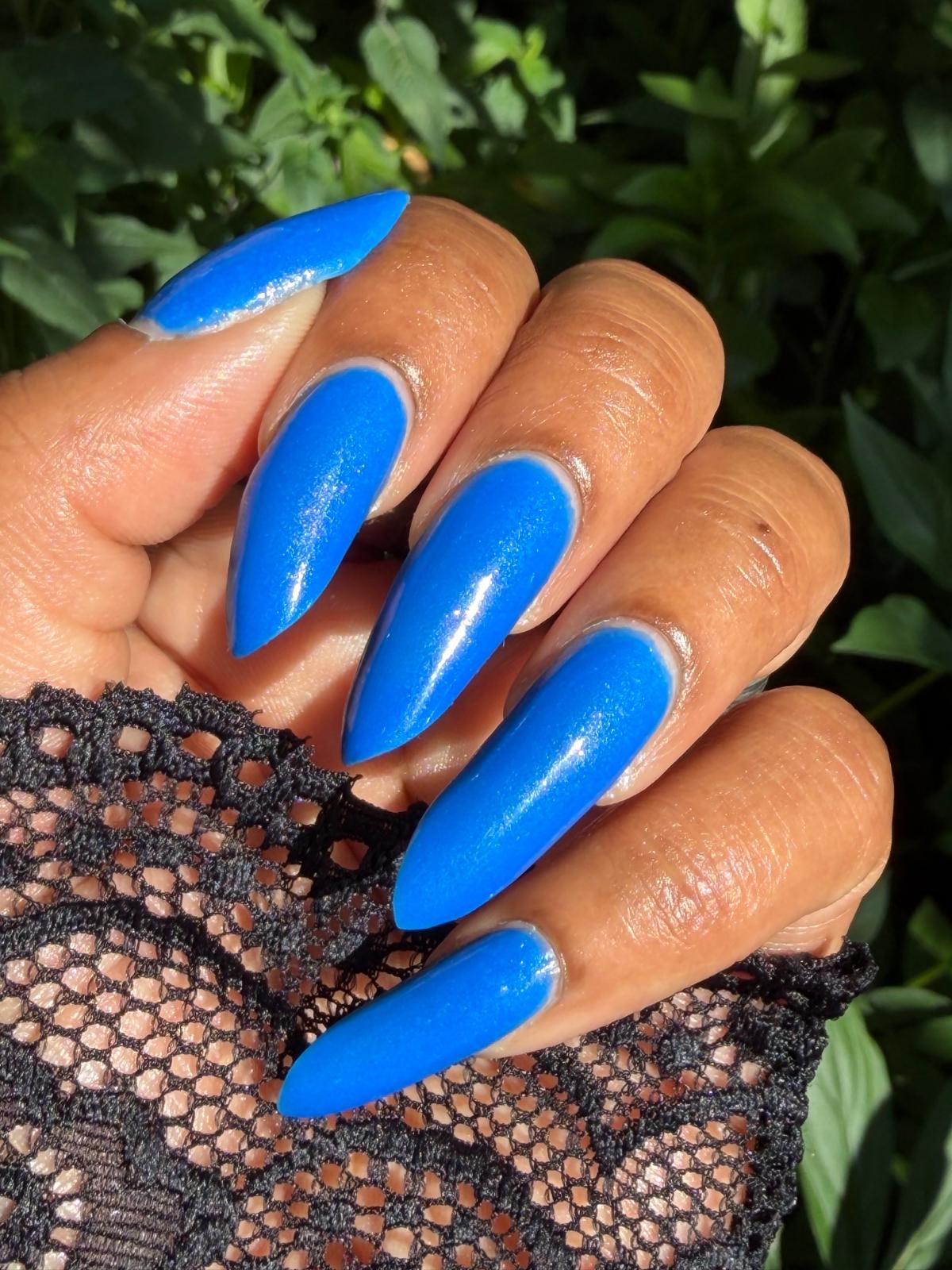 A close-up of a hand with long, glossy, bright blue stiletto-shaped nails, wearing a black lace glove or cuff, with green foliage in the background.