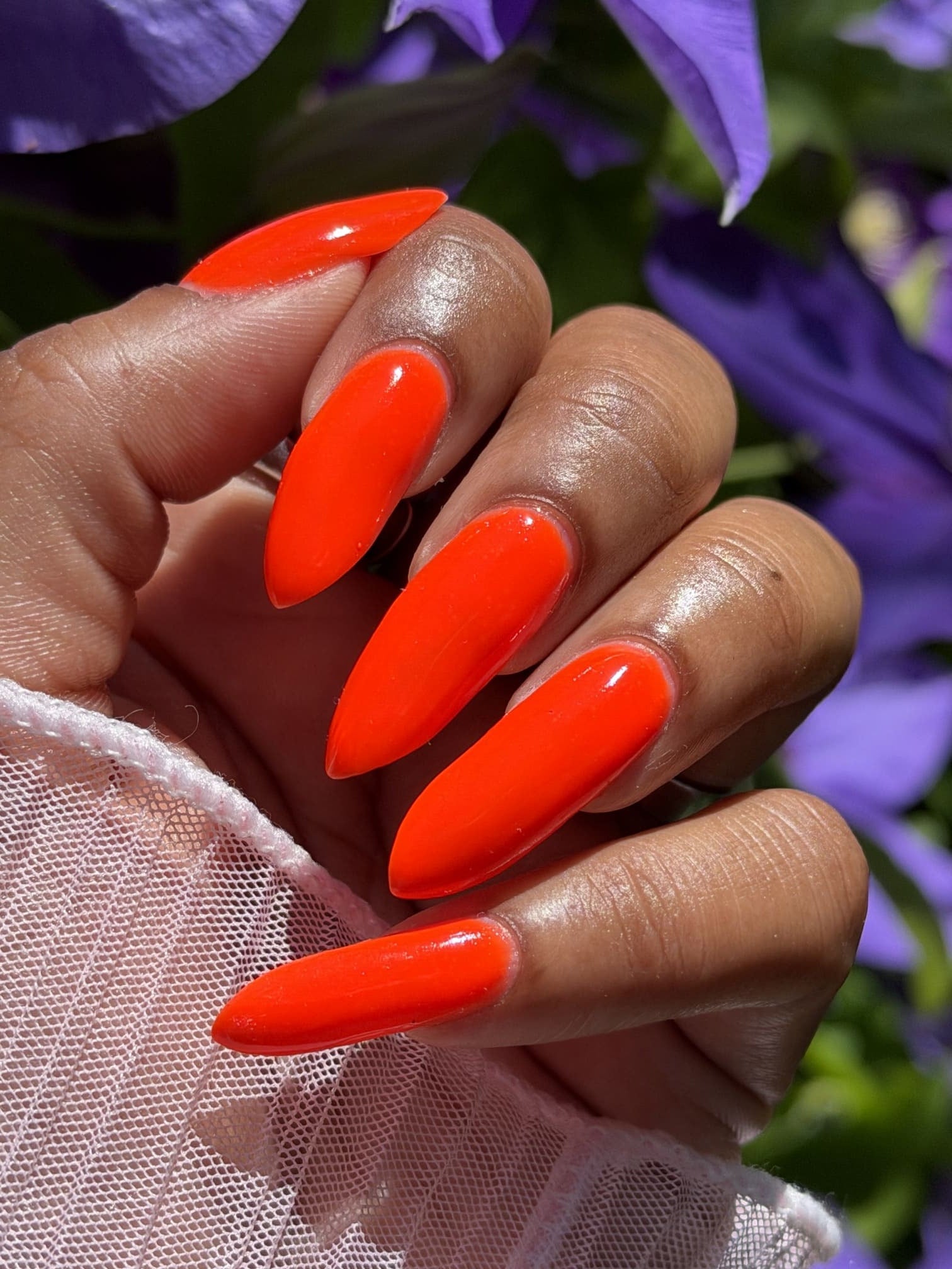 A close-up of a hand with long, glossy, bright neon orange almond-shaped nails, set against a soft-focus background of purple flowers and green leaves.