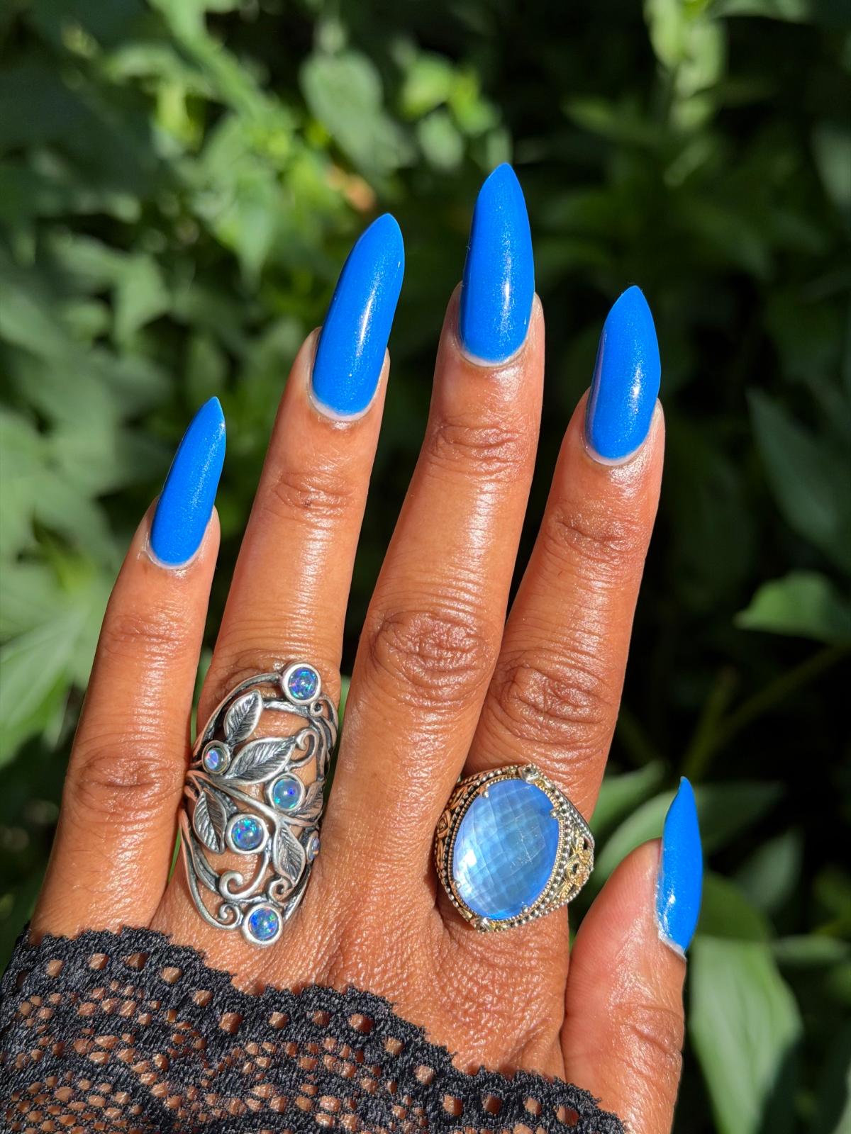 A close-up of a hand with bright blue manicured nails, wearing two rings: a large silver filigree ring with blue stones on the middle finger and a gold ring with a round light blue cabochon on the ring finger, set against a blurred green foliage background.