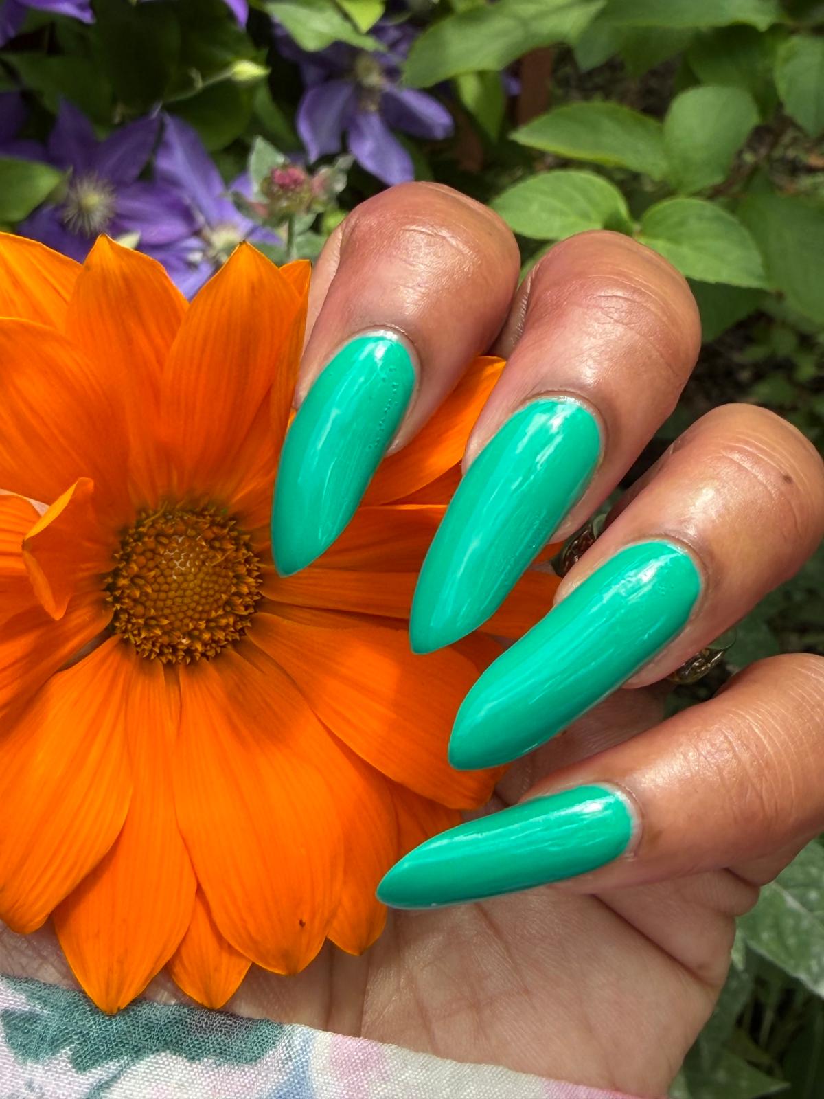 A close-up of a hand with long, glossy mint green stiletto-shaped nails, holding a bright orange flower against a background of green foliage and purple flowers.