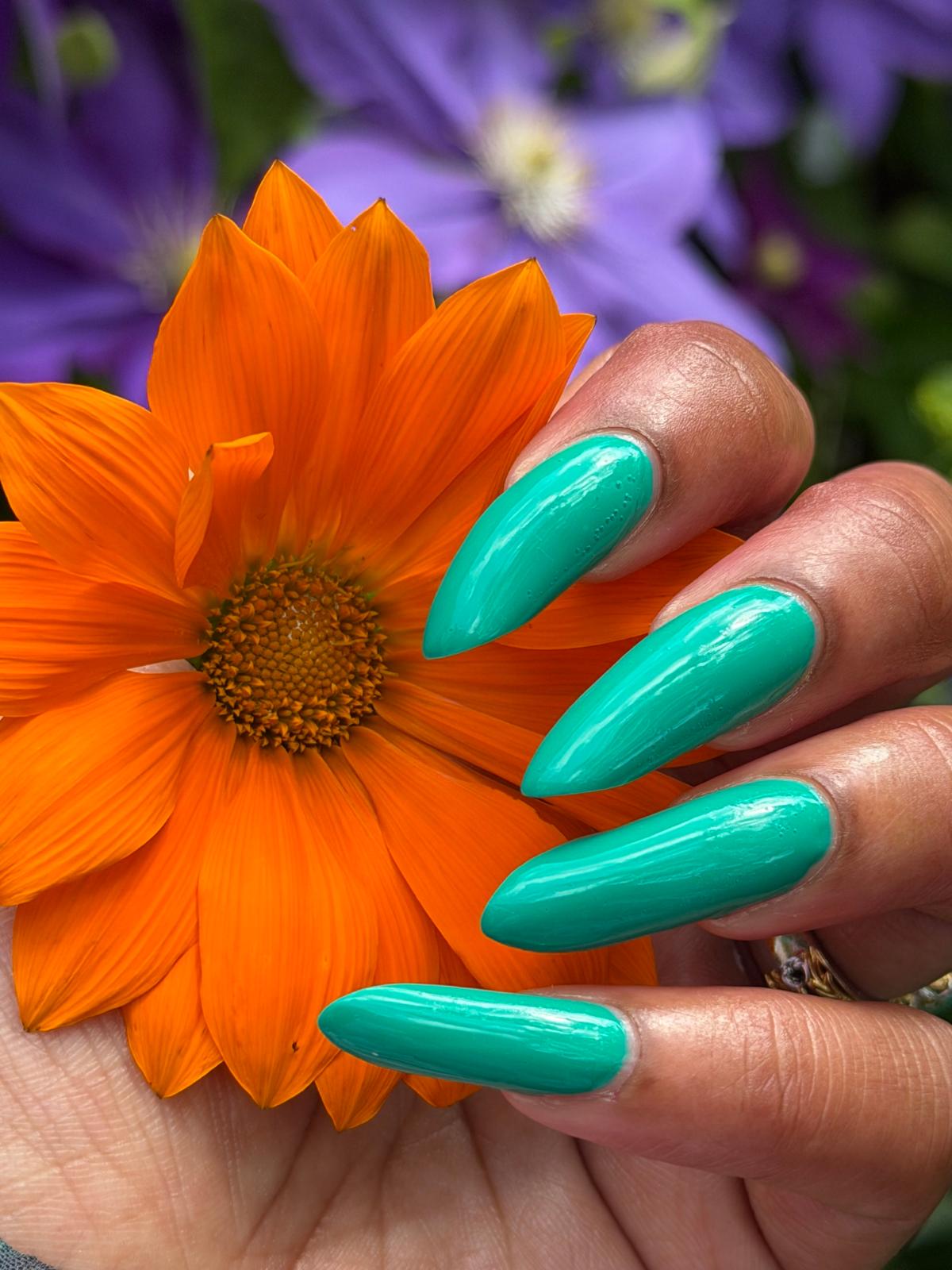 A close-up of a hand with long, glossy teal manicured nails holding a bright orange flower, with blurred purple flowers in the background.