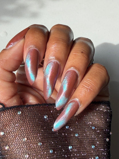 A close-up of a hand with long stiletto-shaped nails painted in a shimmery iridescent pink and light blue holographic finish, next to a sheer black mesh sleeve with rhinestones.
