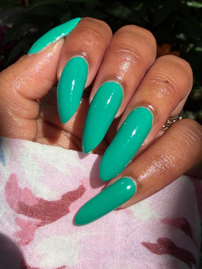 A close-up of a hand with long, glossy almond-shaped nails painted in a bright mint green color, resting on a pink and white patterned fabric with dark green foliage in the background.