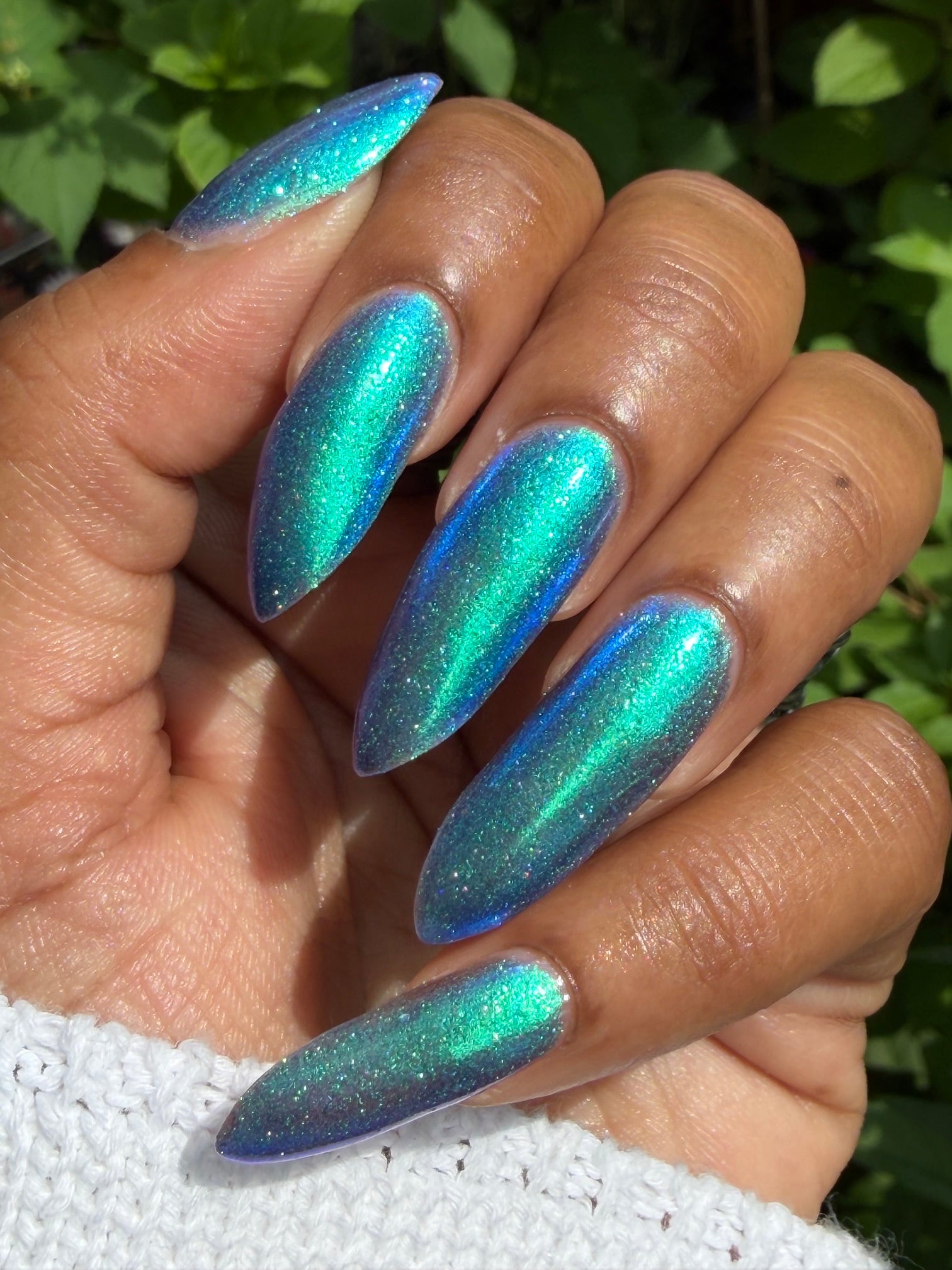 A close-up of a hand with long, stiletto-shaped nails painted in a shimmering holographic teal and blue color that shifts with the light, set against a background of green foliage and a white knit sleeve.