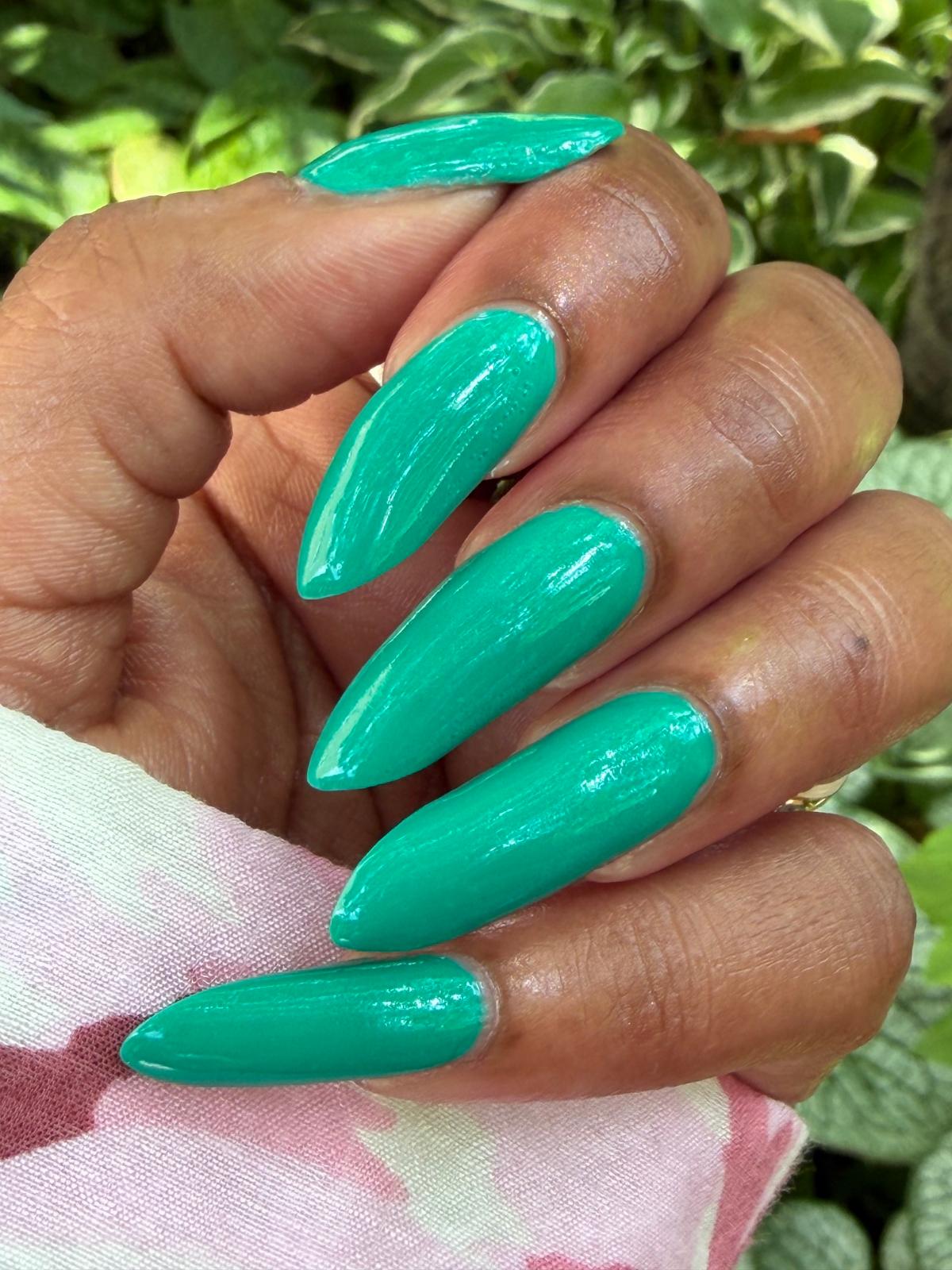 A close-up of a hand with long, glossy, almond-shaped nails painted in a bright mint green color, set against a background of green foliage and a pink patterned fabric.