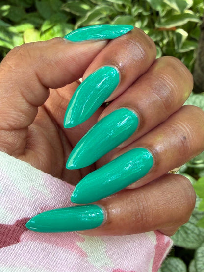 A close-up of a hand with long, glossy, almond-shaped nails painted in a bright mint green color, set against a background of green foliage and a pink patterned fabric.