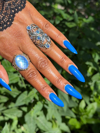 A close-up of a hand with long, bright blue manicured nails, wearing two silver rings: one with a large oval blue stone and another with smaller blue stones, set against a blurred green foliage background.