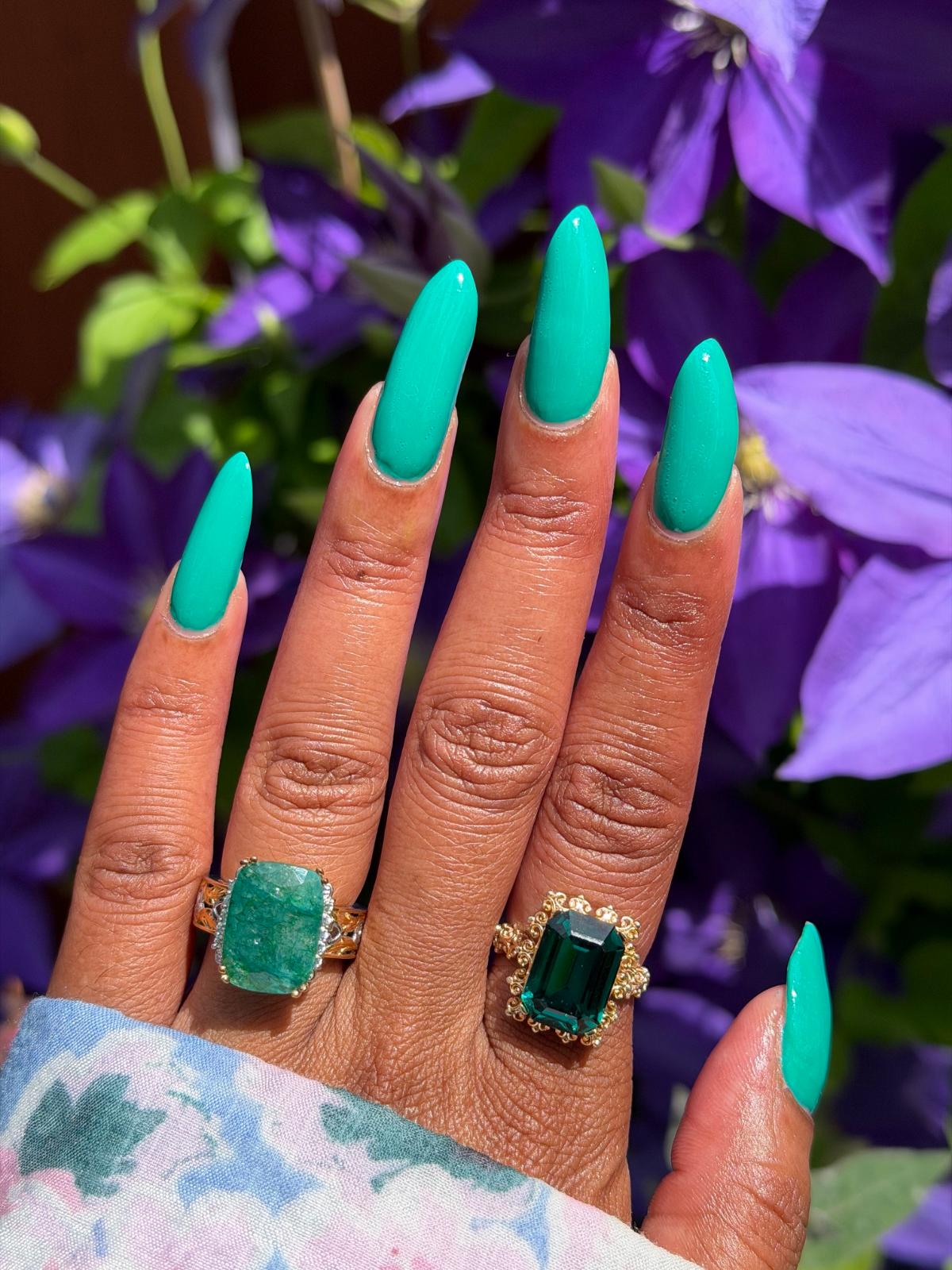 A close-up of a hand with long, glossy emerald green manicured nails, wearing two gold rings with large green emerald-cut gemstones, set against a background of purple flowers.