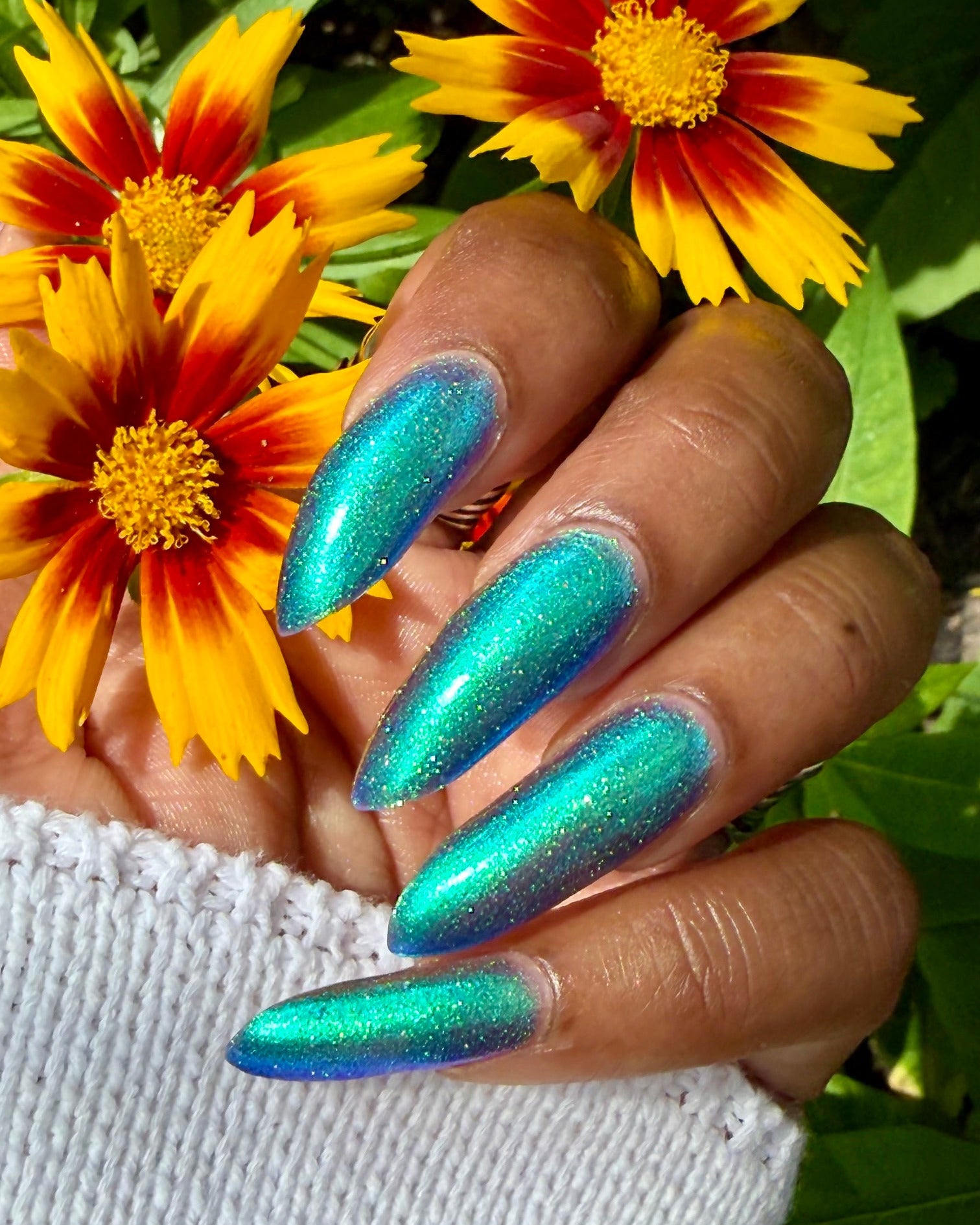 A close-up of a hand with long, almond-shaped nails painted in a shimmering teal and blue holographic nail polish, posed in front of yellow and orange flowers with green leaves.