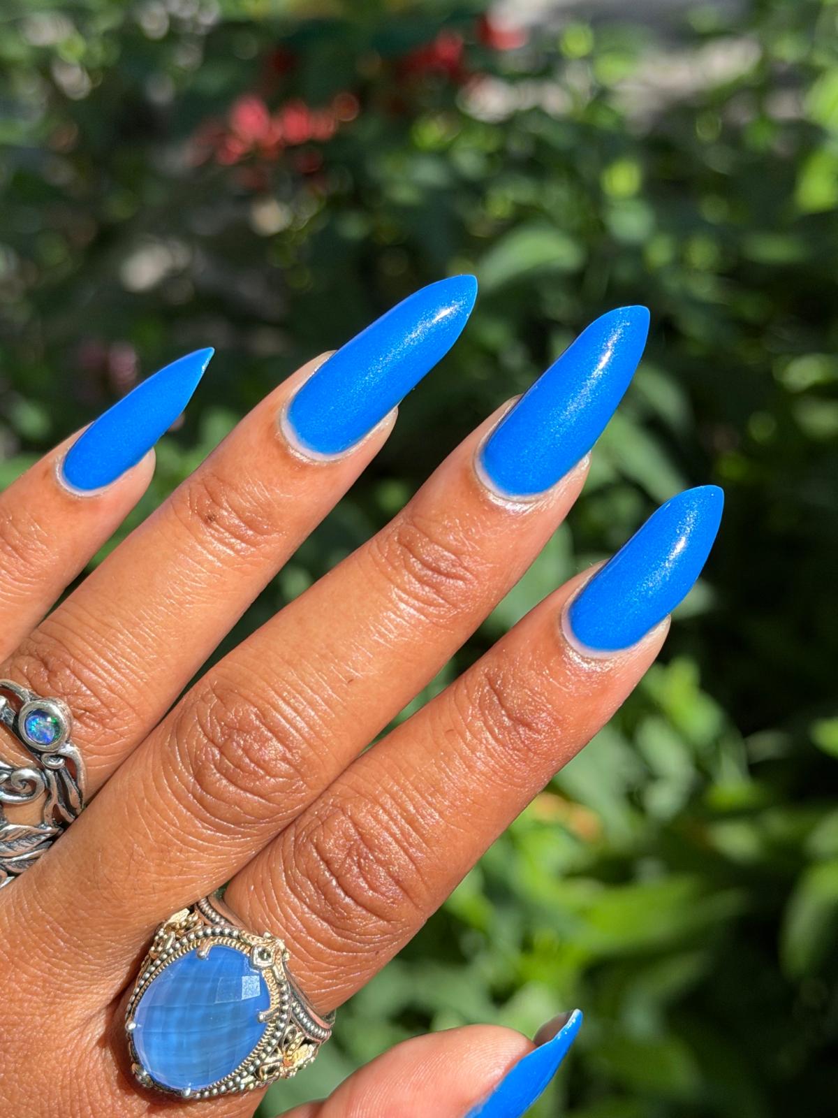 A close-up of a hand with long, glossy bright blue stiletto-shaped press-on nails, wearing silver rings, set against a blurred green garden background.