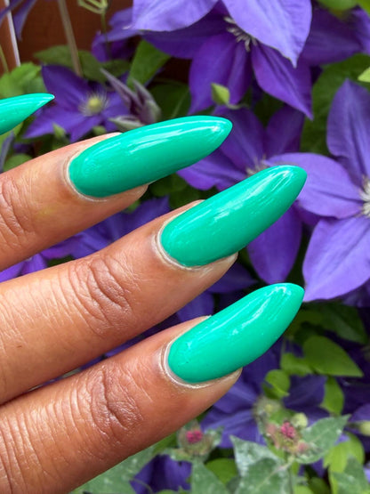 A close-up of a hand with long, glossy, almond-shaped nails painted in a bright mint green or turquoise shade, set against a background of purple clematis flowers.