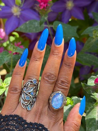 A close-up of a hand with long, bright blue manicured nails, wearing two rings: a large silver filigree ring with blue stones on the middle finger and an oval silver ring with a blue moonstone on the ring finger, set against a blurred background of purple flowers and green leaves.