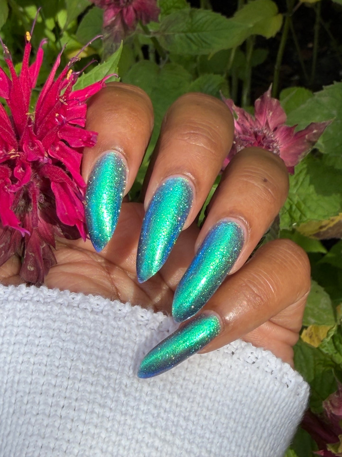 A close-up of a hand with long, almond-shaped nails painted in a shimmering iridescent teal and blue glitter nail polish, styled with a white knit sleeve against a background of green leaves and pink flowers.