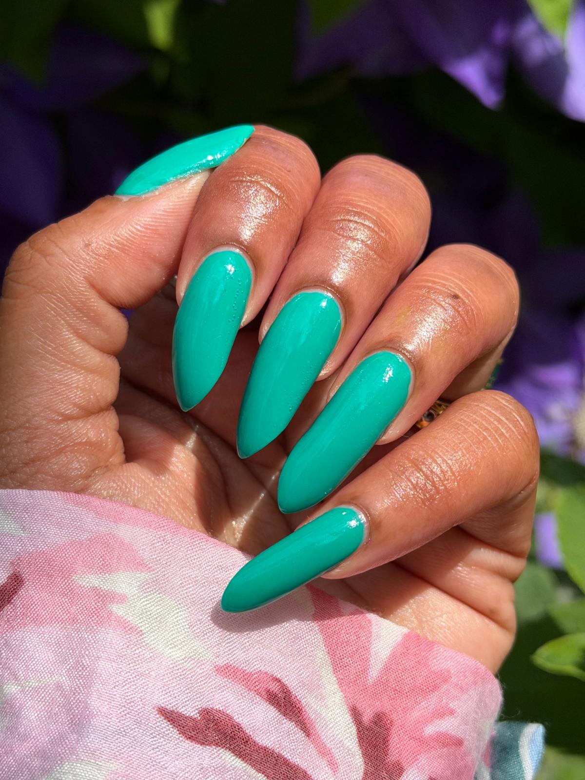 A close-up of a hand with long, almond-shaped nails painted in a glossy bright turquoise green, resting against a pink floral fabric with blurred green leaves and purple flowers in the background.