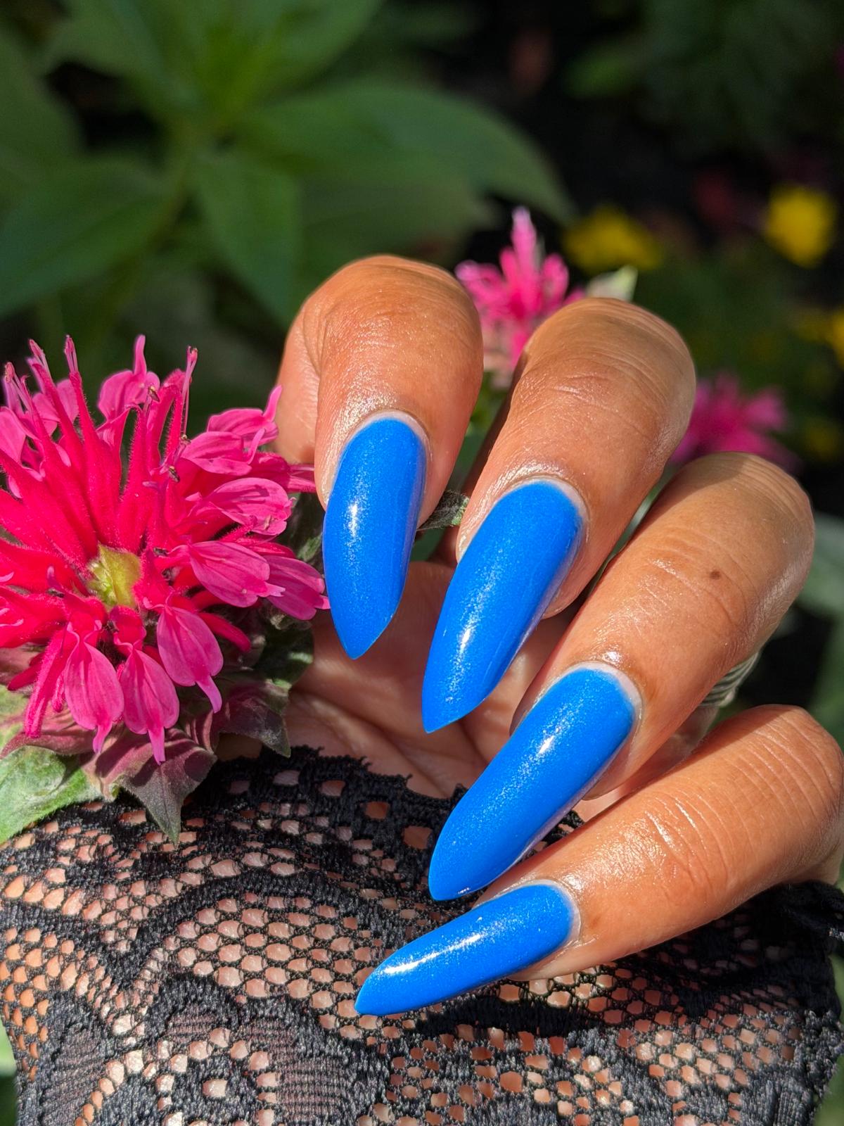 A close-up of a hand with long, glossy bright blue stiletto-shaped nails, resting on a black lace sleeve with pink flowers and green foliage in the background.