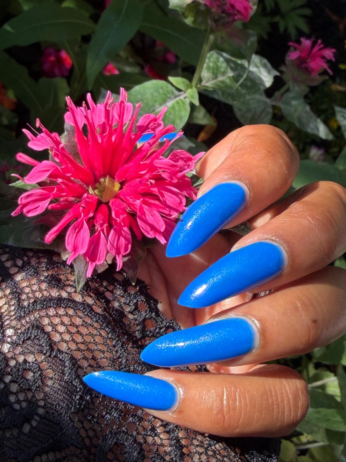 A close-up of a hand with long, glossy bright blue stiletto-shaped nails, holding a vibrant pink flower against a background of green foliage.