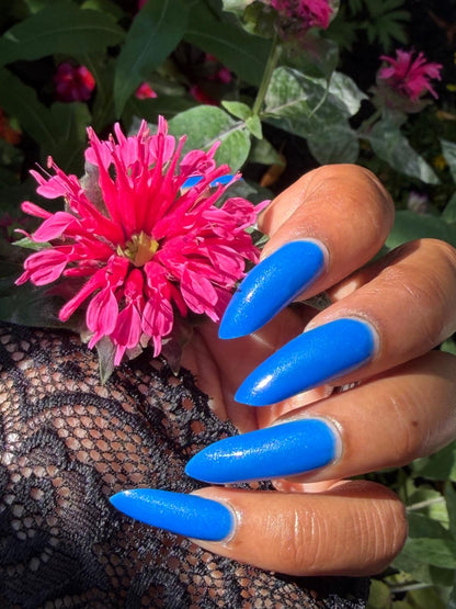 A close-up of a hand with long, glossy bright blue stiletto-shaped nails, holding a vibrant pink flower against a background of green foliage.