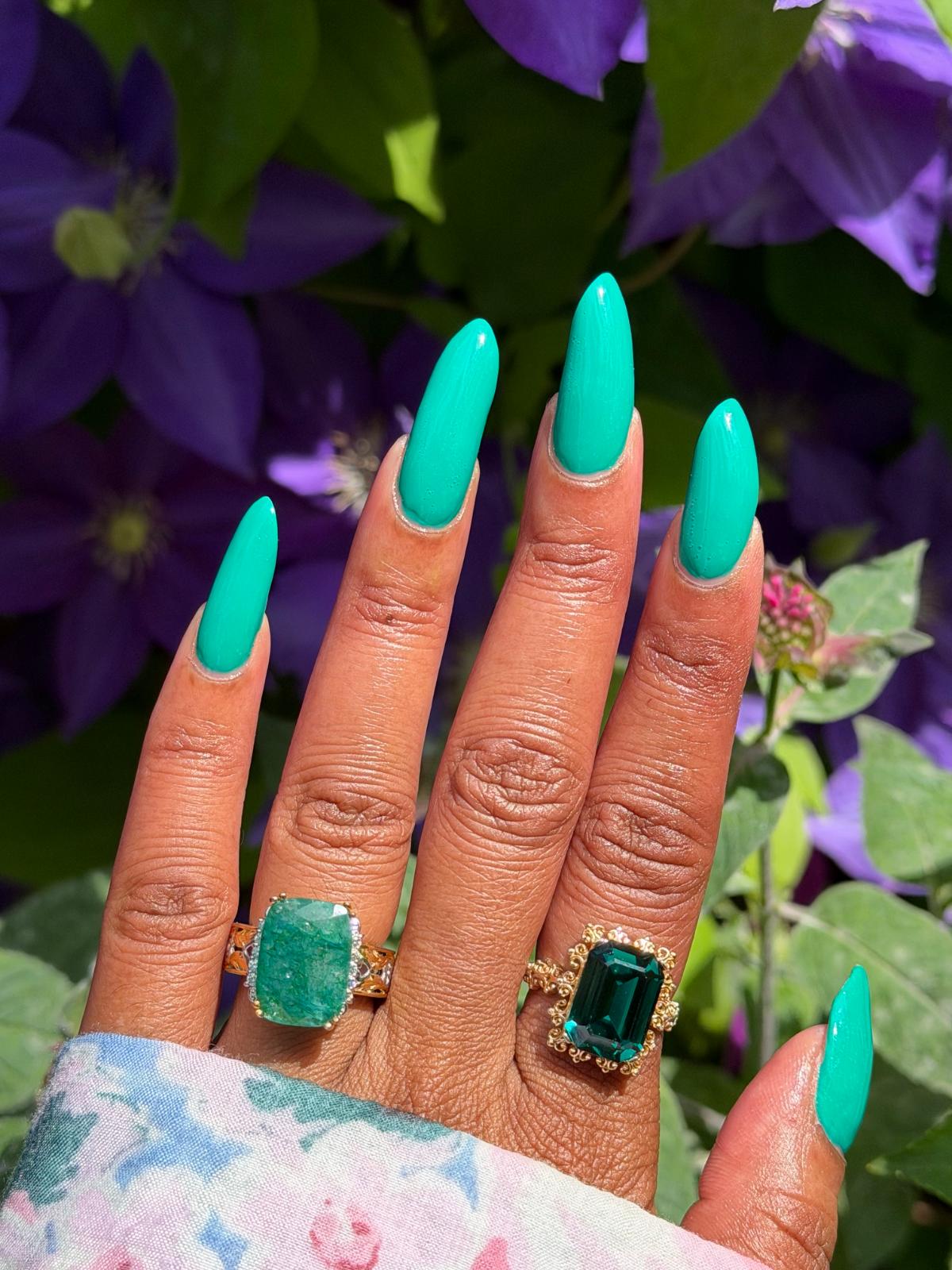 A close-up of a hand with long, glossy turquoise manicured nails, wearing two gold rings with large emerald-cut green gemstones, set against a background of purple flowers and green leaves.