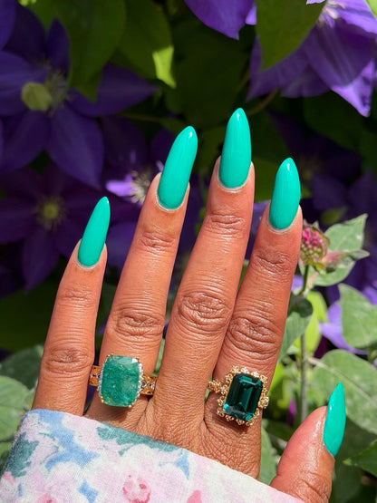 A close-up of a hand with long, glossy turquoise manicured nails, wearing two gold rings with large emerald-cut green gemstones, set against a background of purple flowers and green leaves.