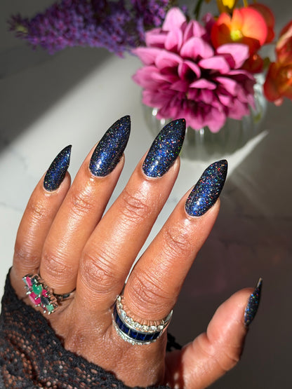 A close-up of a hand with long, almond-shaped nails painted in a dark navy glitter nail polish, styled with rings and a black lace sleeve, with blurred pink and purple flowers in the background.