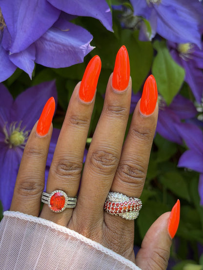 A close-up of a hand with long, bright orange manicured nails, wearing two silver rings with red and clear stones, set against a background of purple flowers and green leaves.