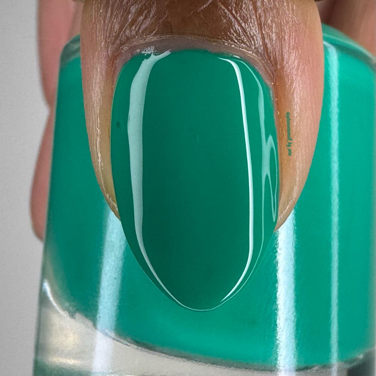 A close-up of a fingernail painted with a glossy bright teal-green nail polish, with the matching polish visible in the bottle behind it.