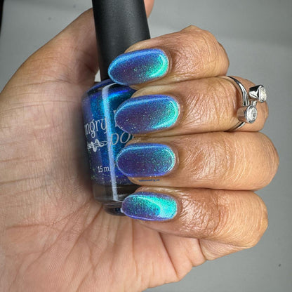 A hand with nails painted in a shimmering blue and teal nail polish, holding a bottle of the same nail polish. The hand also wears a silver ring with two clear stones.