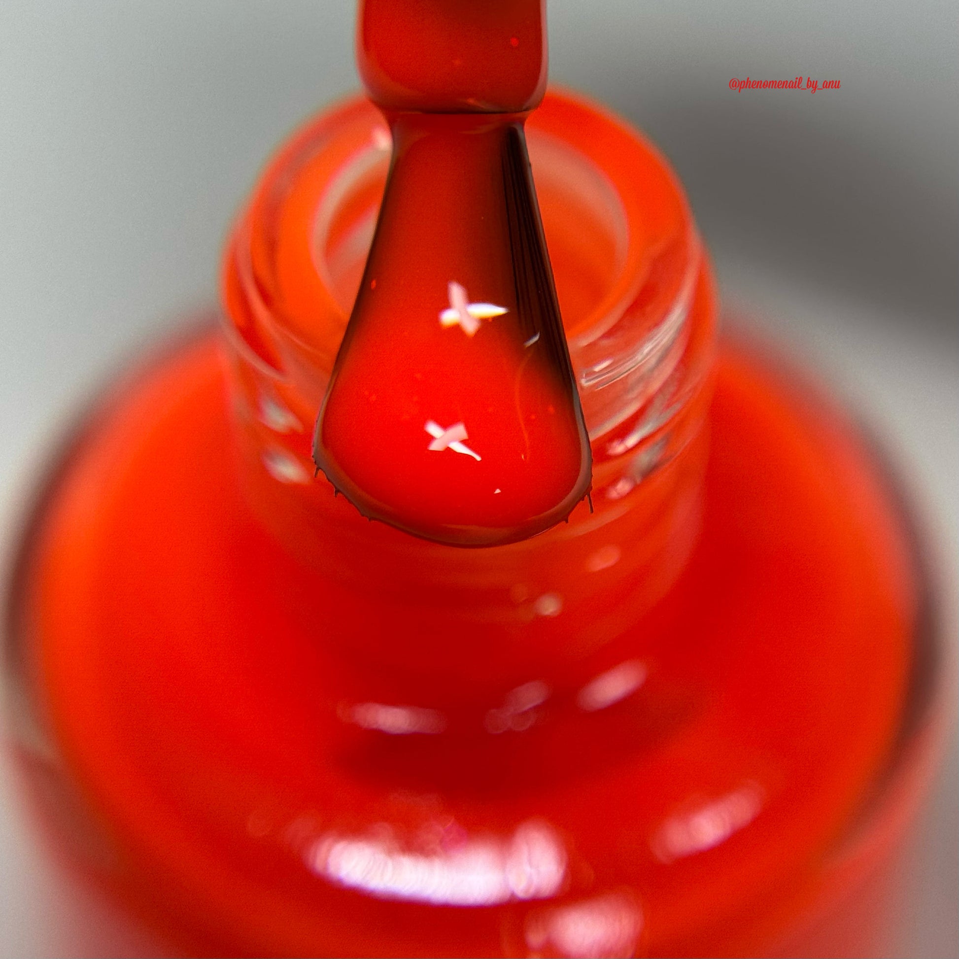 A close-up shot of a glossy red nail polish brush coated with vibrant red polish, positioned above a matching glass bottle of red nail polish against a soft gray background.