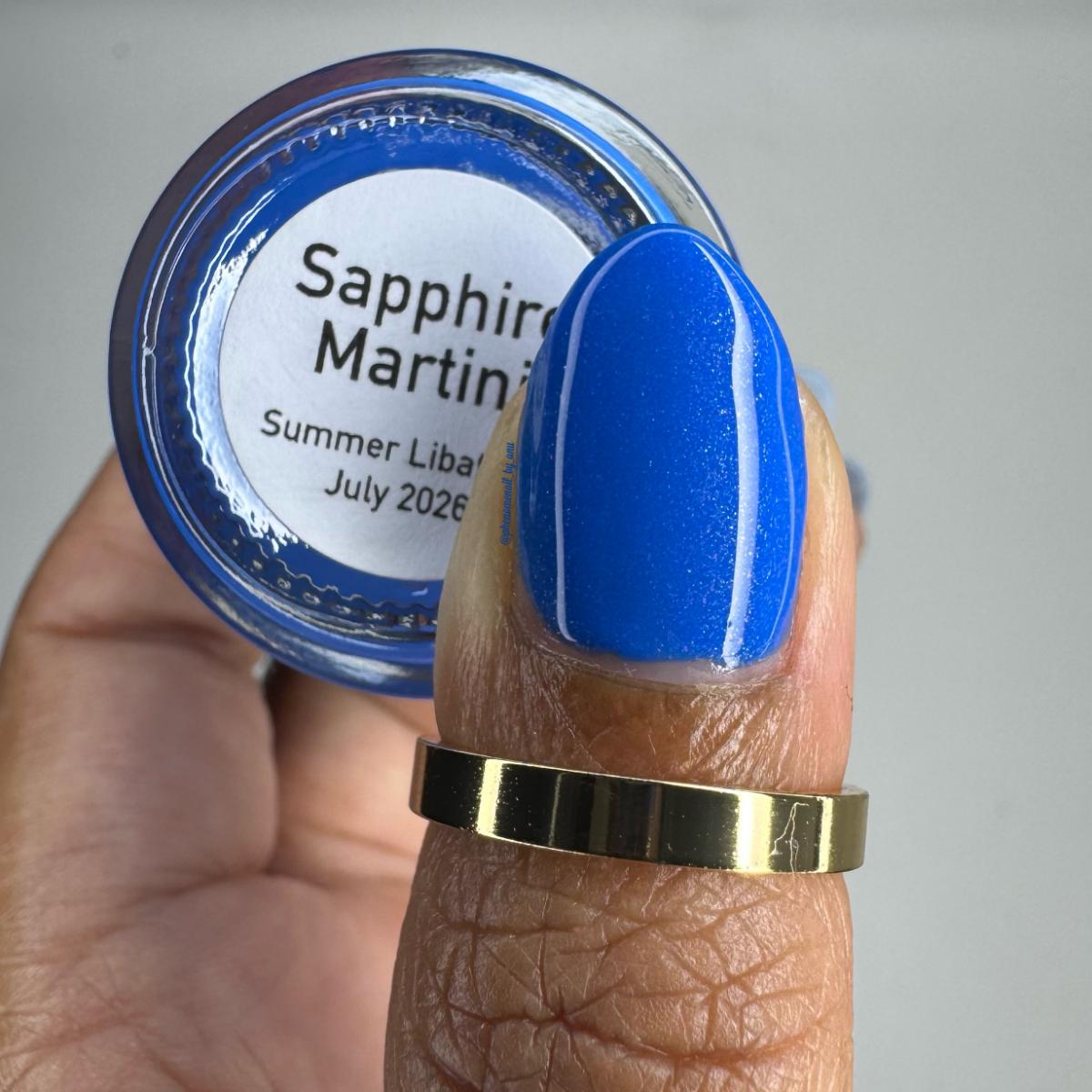 A close-up of a hand holding a small round jar labeled 'Sapphire Martin' with a bright blue nail polish swatch on the fingernail. The jar lid shows the brand name 'Sapphire Martin' and the text 'Summer Liba.
