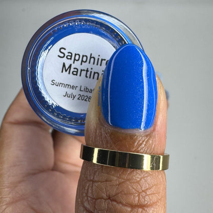 A close-up of a hand holding a small round jar labeled 'Sapphire Martin' with a bright blue nail polish swatch on the fingernail. The jar lid shows the brand name 'Sapphire Martin' and the text 'Summer Liba.