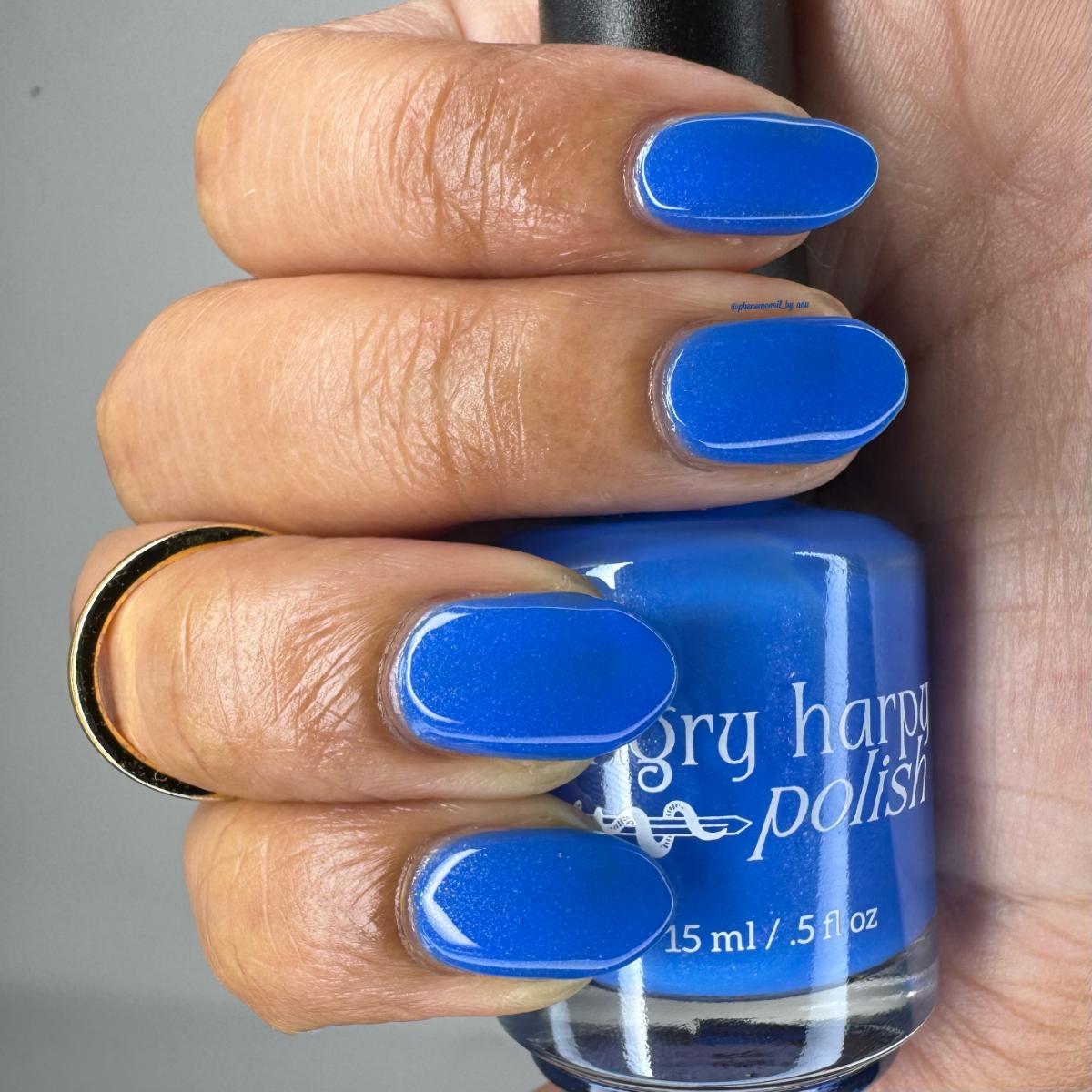 A close-up of a hand holding a bottle of blue nail polish, with the nails painted in a matching bright medium blue shade. The bottle label reads 'Gry harpy polish'.