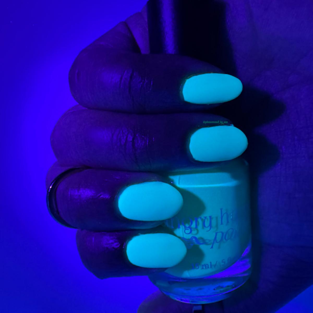 A hand holding a bottle of nail polish under blue light, showing the nails painted in a bright fluorescent light blue shade that glows under UV. The bottle label has purple text but is partially obscured.