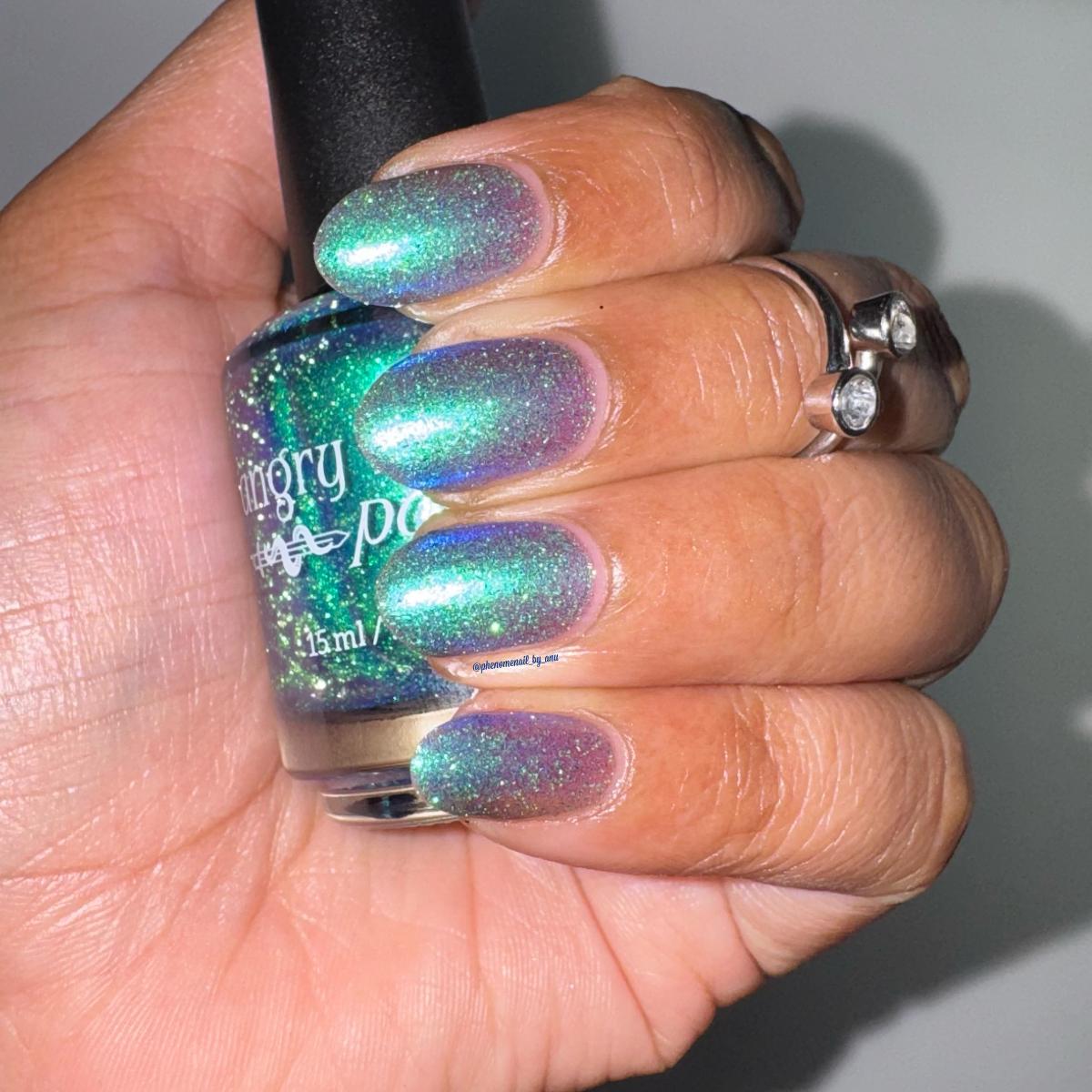 A hand with manicured nails painted in a green-to-purple shimmer nail polish, holding a bottle of the same glittery nail polish under flash lighting.