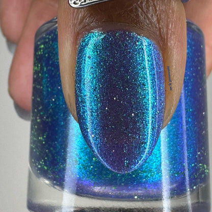 A close-up of a fingernail painted with a shimmering blue-to-purple nail polish, with the clear bottle of matching polish visible behind it.