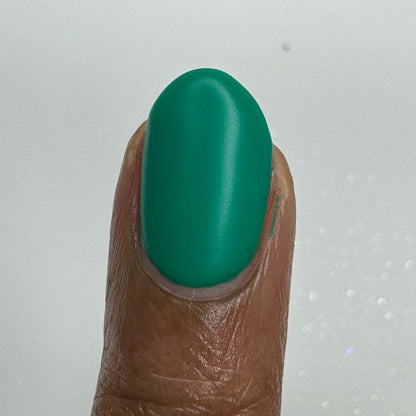 A close-up of a fingernail painted with a solid, glossy emerald green polish against a light gray background.