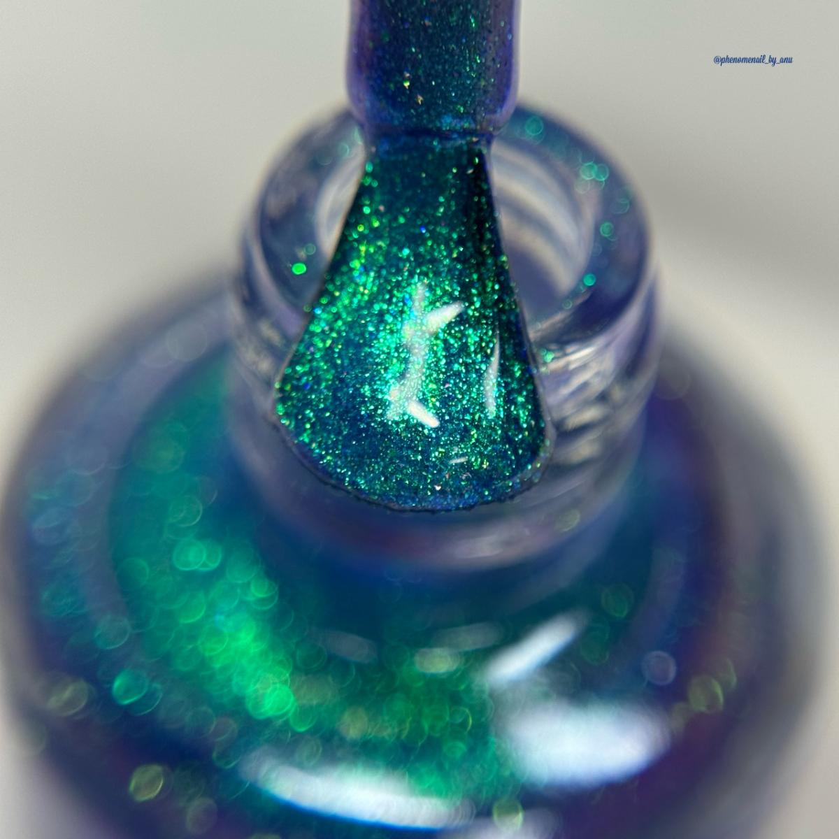 A close-up of a nail polish bottle with the brush covered with a layer of glittery green and blue shimmer polish.