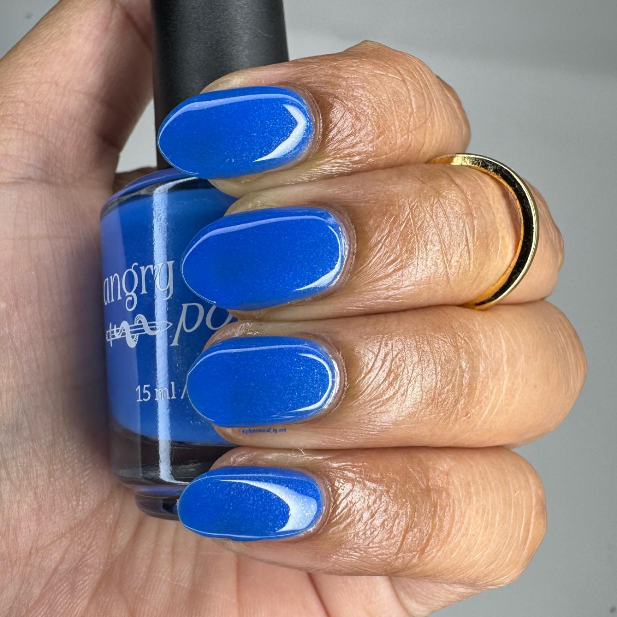 A close-up of a hand with fingernails painted in a bright royal blue polish, holding a matching blue nail polish bottle. The hand also wears a gold ring, and the background is a plain light gray.