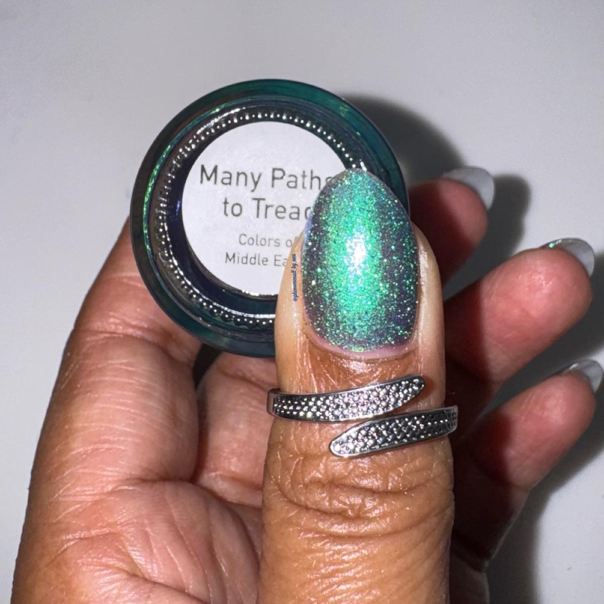 A hand holding a nail polish bottle, with the thumb painted in a sparkling green-to-blue metallic polish under flash lighting. The bottle label reads 'Many Paths to Tread' and 'Colors of Middle Earth.