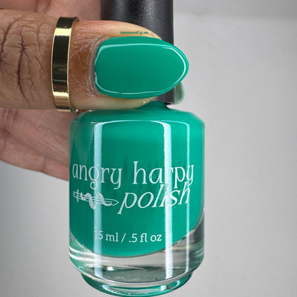 A hand holding a bottle of Angry Harpy Polish nail lacquer, showing the teal-green polish color applied to a fingernail against a plain light background.
