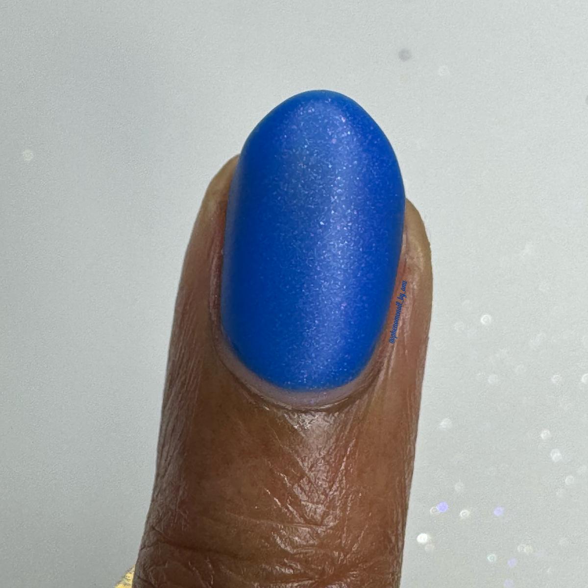 A close-up of a fingernail painted with a vibrant, shimmery royal blue nail polish, shown against a light gray background with subtle glitter.