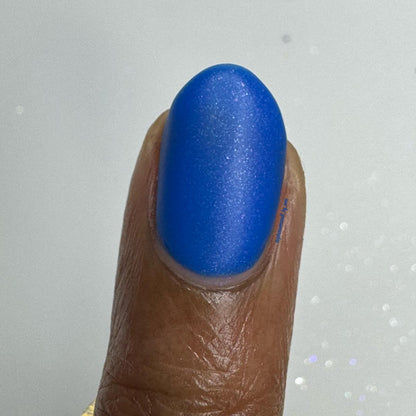 A close-up of a fingernail painted with a vibrant, shimmery royal blue nail polish, shown against a light gray background with subtle glitter.