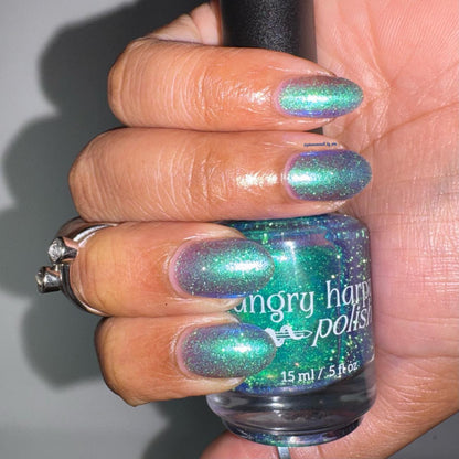 A hand holding a bottle of nail polish with matching iridescent green-blue nails painted on, against a plain gray background.