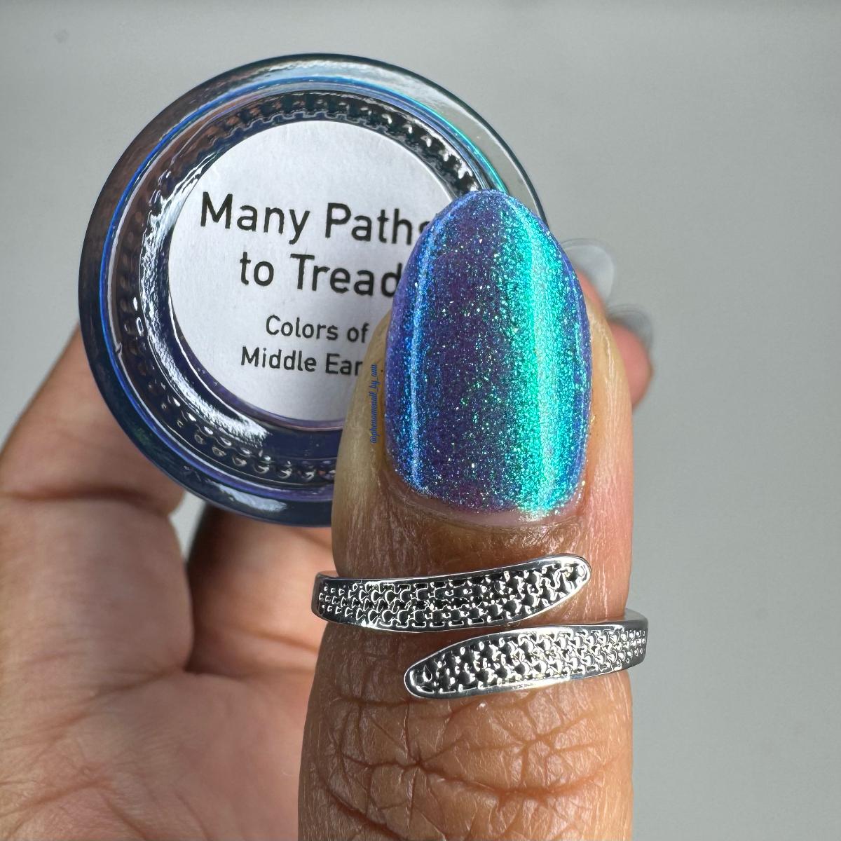 A close-up of a hand holding a nail polish bottle with thumbnail painted with the same shimmering blue-purple-green color. The bottle label reads 'Many Paths to Tread' and 'Colors of Middle Earth".