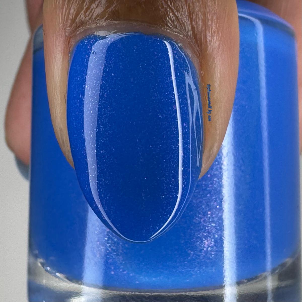 A close-up of a fingernail painted with a vivid medium blue nail polish, positioned in front of a matching blue nail polish bottle.